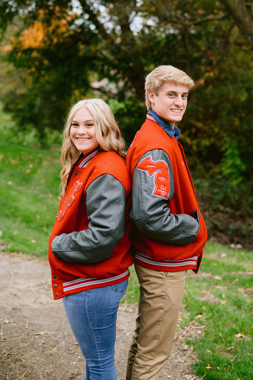Guy and girl wearing matching Letterman jackets and standing back-to-back as they smile and pose for their senior wedding shoot with Mary Shelton Media