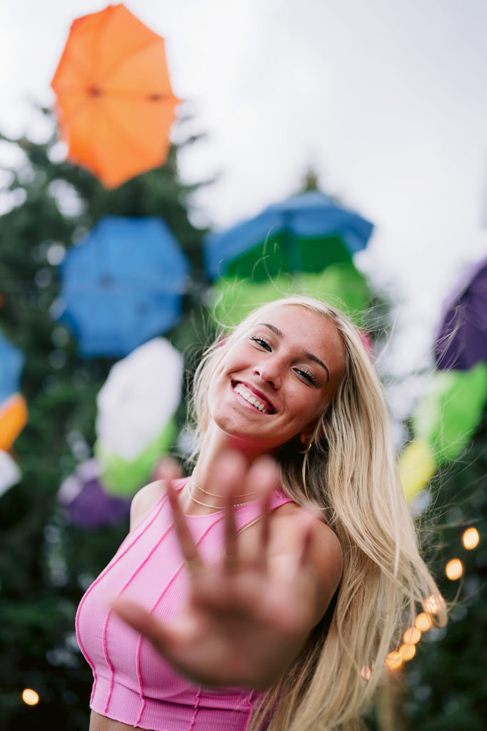 Elegant girl holding her hand up and smiling at the camera with colorful umbrellas and fairy lights behind her during their shoot with Mary Shelton Media