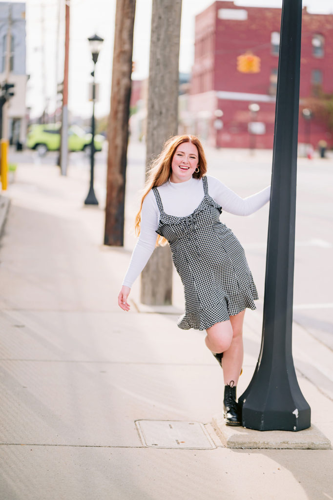 Girl smiles for the camera as she holds on to a lamp post on the sidewalk during their senior shoot with Michigan photographer Mary Shelton