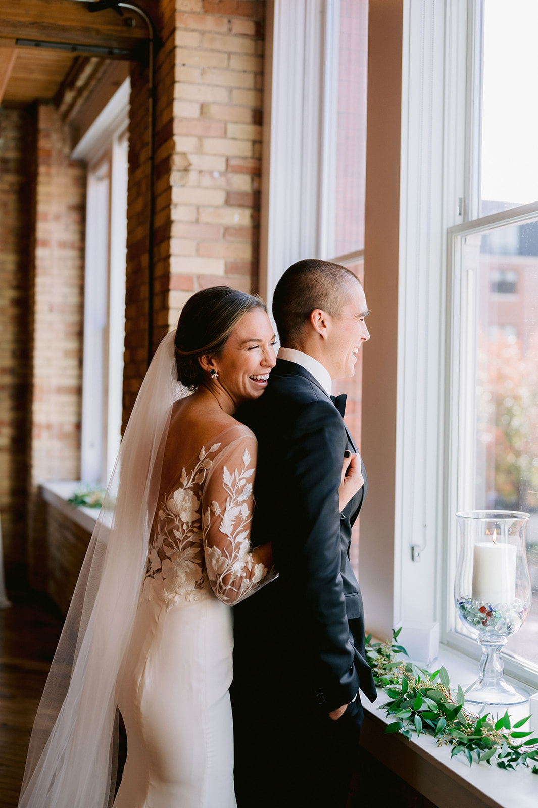 Bride smiles as she hugs groom from behind as he looks out the window, captured by Mary Shelton Media